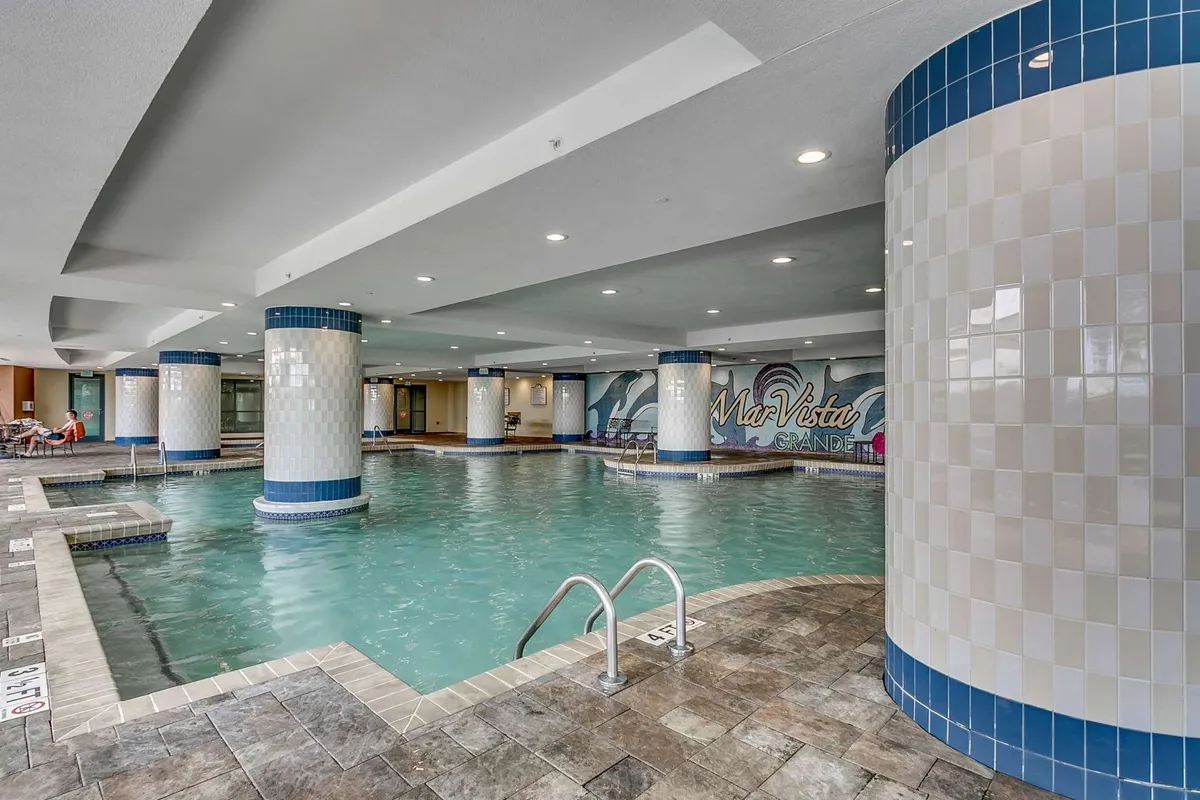 A spacious indoor swimming pool area featuring decorative tile columns, a vibrant mural along the wall, several lounging chairs, and ample natural light. The pool is calm and inviting, ideal for both swimming and relaxing.