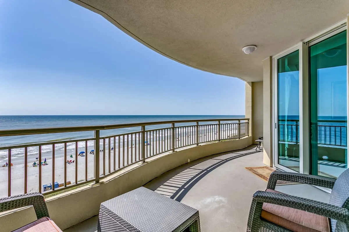 A private oceanfront balcony furnished with comfortable outdoor seating, overlooking the serene beach and gentle waves. Umbrellas dot the sandy shore below, and the inviting scene hints at both sunrise and sunset moments, perfectly aligning with the section’s references to morning coffee and sunset cocktails.