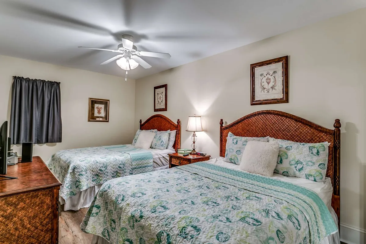 A comfortable guest bedroom with two beds featuring decorative headboards, a nightstand topped with a lamp, and framed artwork adorning the walls. Natural light filters through the curtains, creating a restful, cozy ambiance.