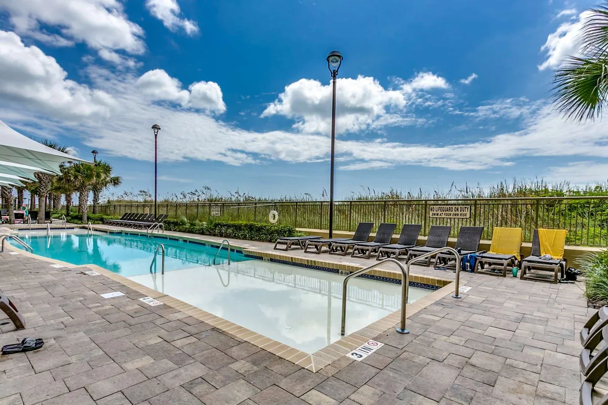 An outdoor swimming pool surrounded by lush greenery, with plenty of lounge chairs arranged for sunbathing and relaxation. The sky is clear, and the scene captures a leisurely resort vibe.
