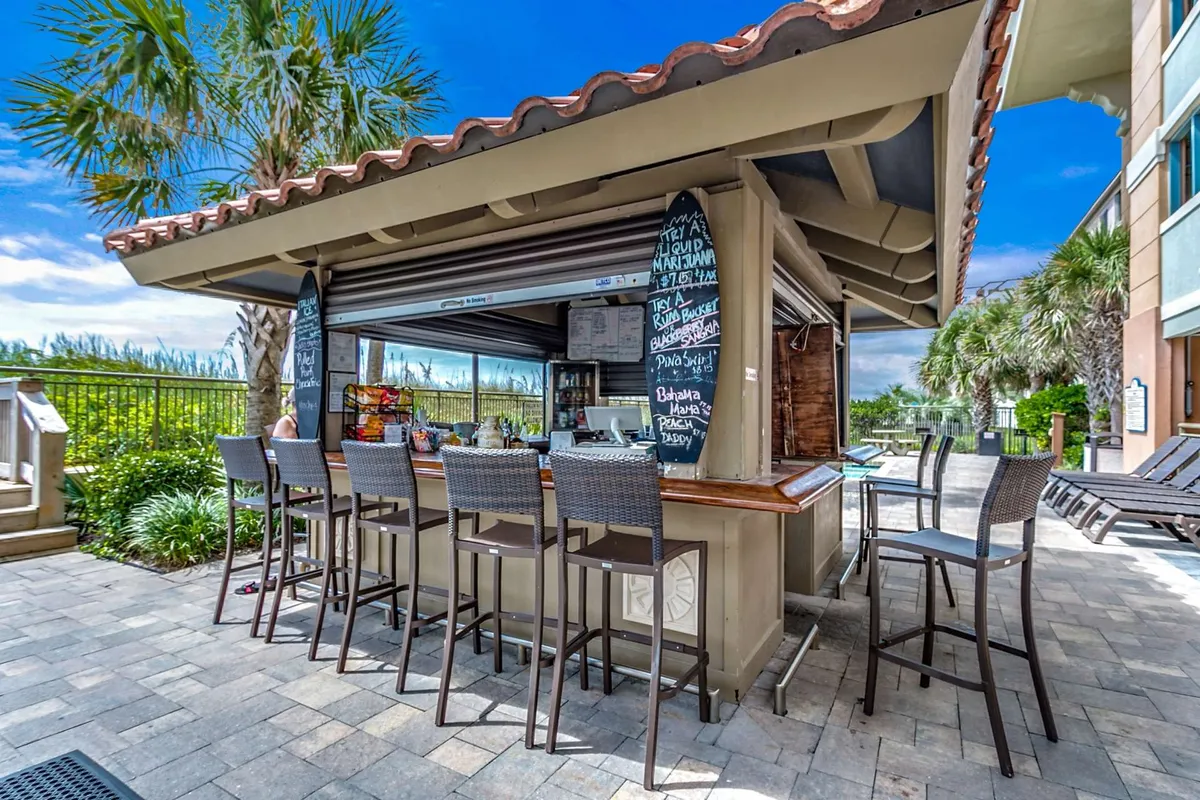This exterior image features an inviting outdoor bar area with bistro chairs, a chalkboard menu, palm trees, and a swimming pool, all basking in natural light. The relaxed, social atmosphere captures guests enjoying drinks, snacks, and poolside lounging.