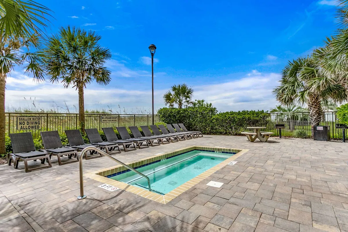 A vibrant outdoor pool area lined with palm trees and lounge chairs, featuring clear blue water and poolside signage. The setting is inviting and lively, ideal for both sunbathing and swimming, and hints at the property’s resort-like amenities for all ages.