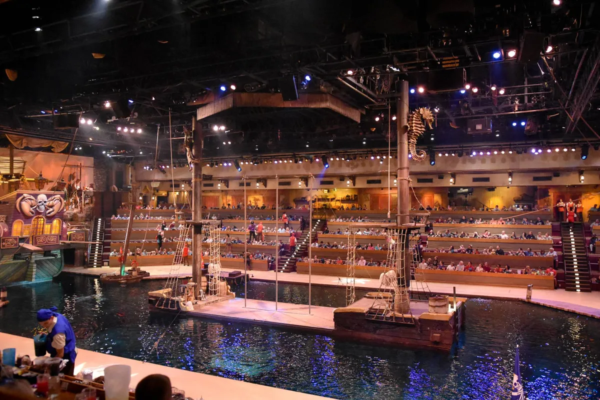 An energetic scene inside a live performance venue featuring a large pirate ship structure as a stage, a water stage for aquatic shows, and rows of seating filled with an engaged audience. The space is vibrant and full of anticipation.