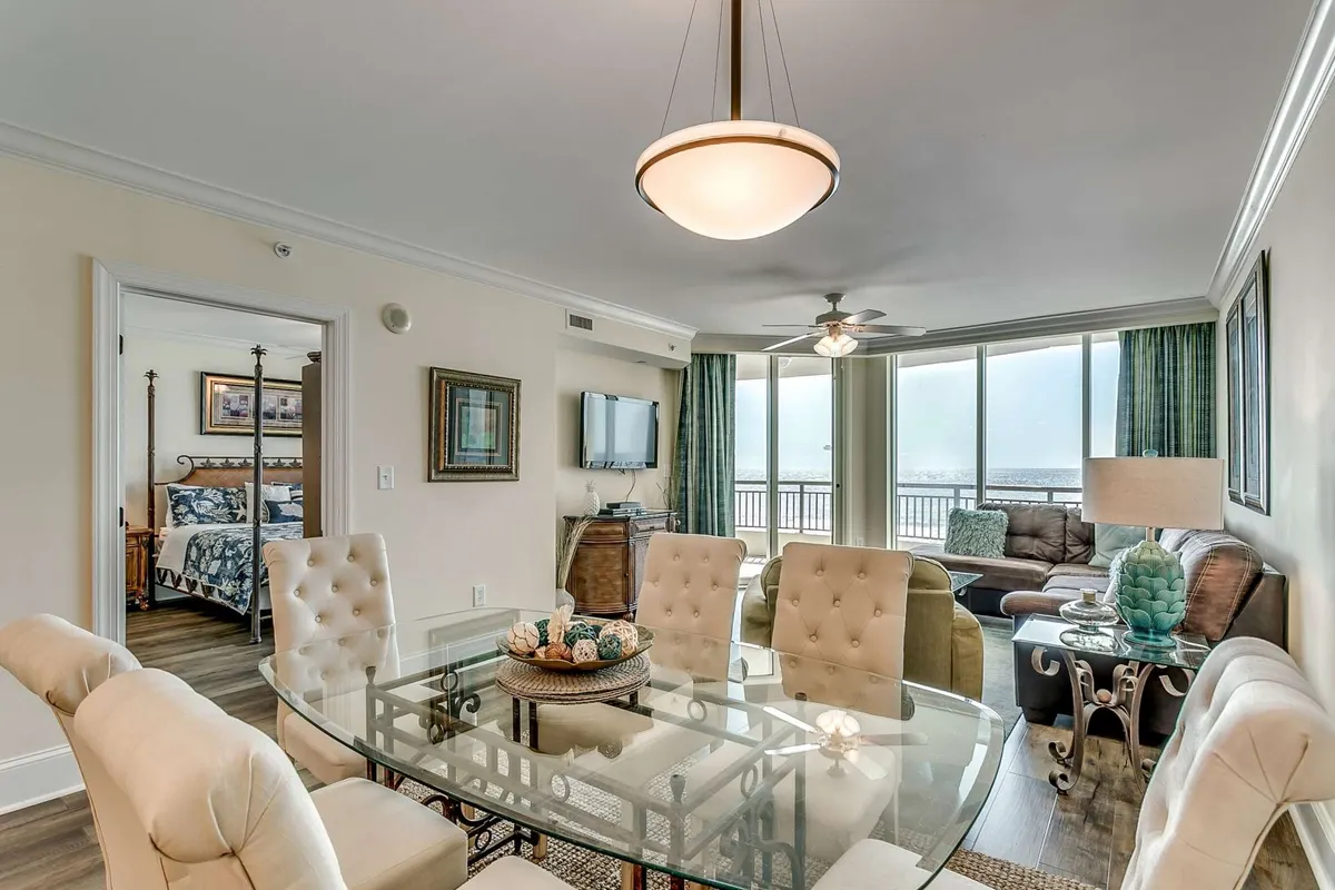 The image showcases a bright, open-concept living and dining area featuring a glass dining table set for family meals, a plush seating area perfect for relaxing, and expansive windows that frame breathtaking beach views. Decorative accents add warmth and a touch of coastal elegance, while natural light fills the space, creating a serene and inviting atmosphere.