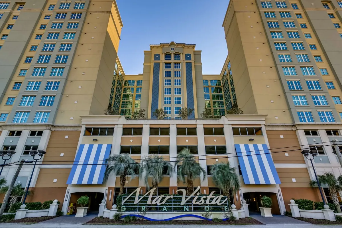 An exterior view of the resort’s tall building with its welcoming entrance sign, striped awning, and lush palm landscaping. The scene communicates the resort’s grandeur and proximity to coastal attractions.