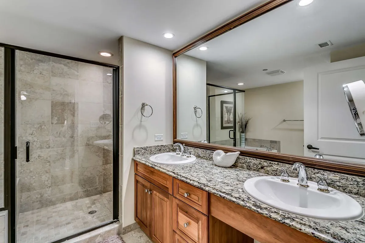 A modern bathroom with a double vanity topped in granite, a large mirror reflecting soft lighting, and a glass-enclosed shower. The space feels serene and functional, ideal for morning routines or unwinding at night.