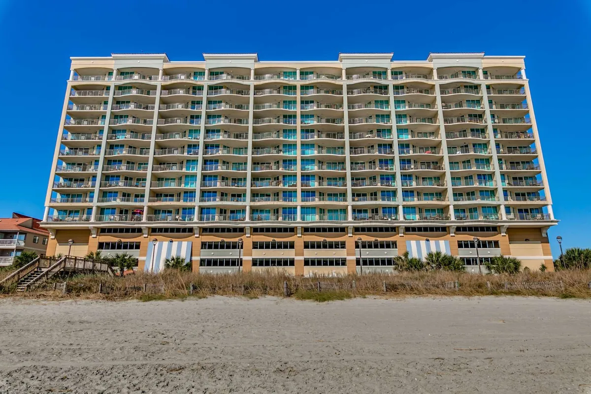 The image features the facade of Mar Vista Grande, showcasing multiple balconies overlooking a lush, vibrant landscape with the beach nearby. The sun illuminates the building, and the surrounding area hints at easy access to the ocean and community amenities.