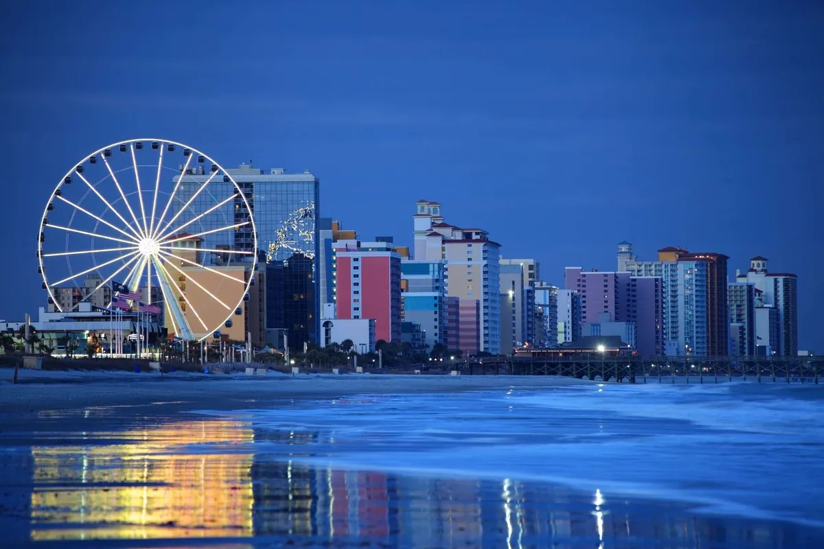 A lively boardwalk scene at sunset with a ferris wheel, colorful buildings, and shimmering ocean reflections. The setting invites evening strolls, amusement rides, and nearby dining, creating an energetic and welcoming atmosphere.