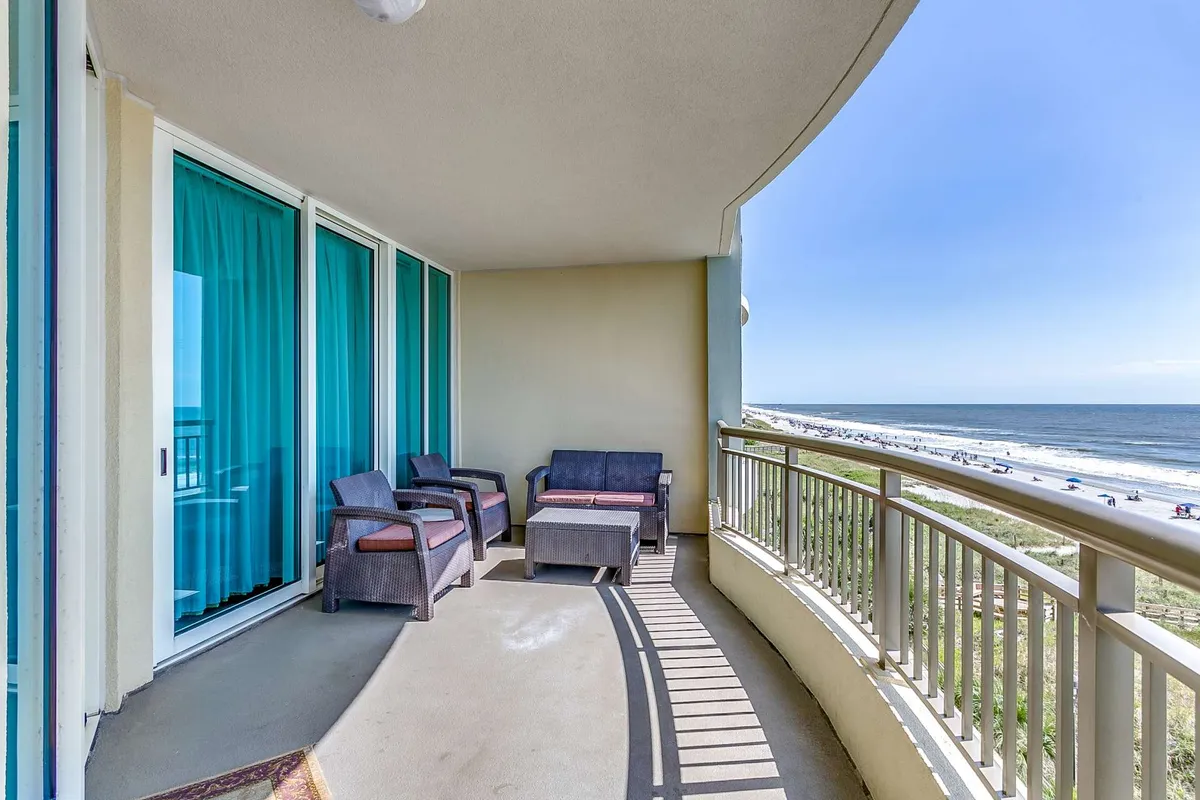 A breathtaking view of the beach as seen from a private balcony, with inviting outdoor seating and a stylish curved railing. The scene is calm and bathed in natural light, suggesting the perfect spot for families to gather and enjoy the ocean breeze and sunset views after a fun-filled day.