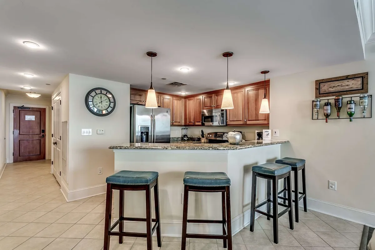 A warm interior shot featuring a granite countertop island with stools, kitchen cabinets, and visible appliances including a dishwasher and coffee maker. The open layout hints at the sociable atmosphere ideal for group meals or entertaining.