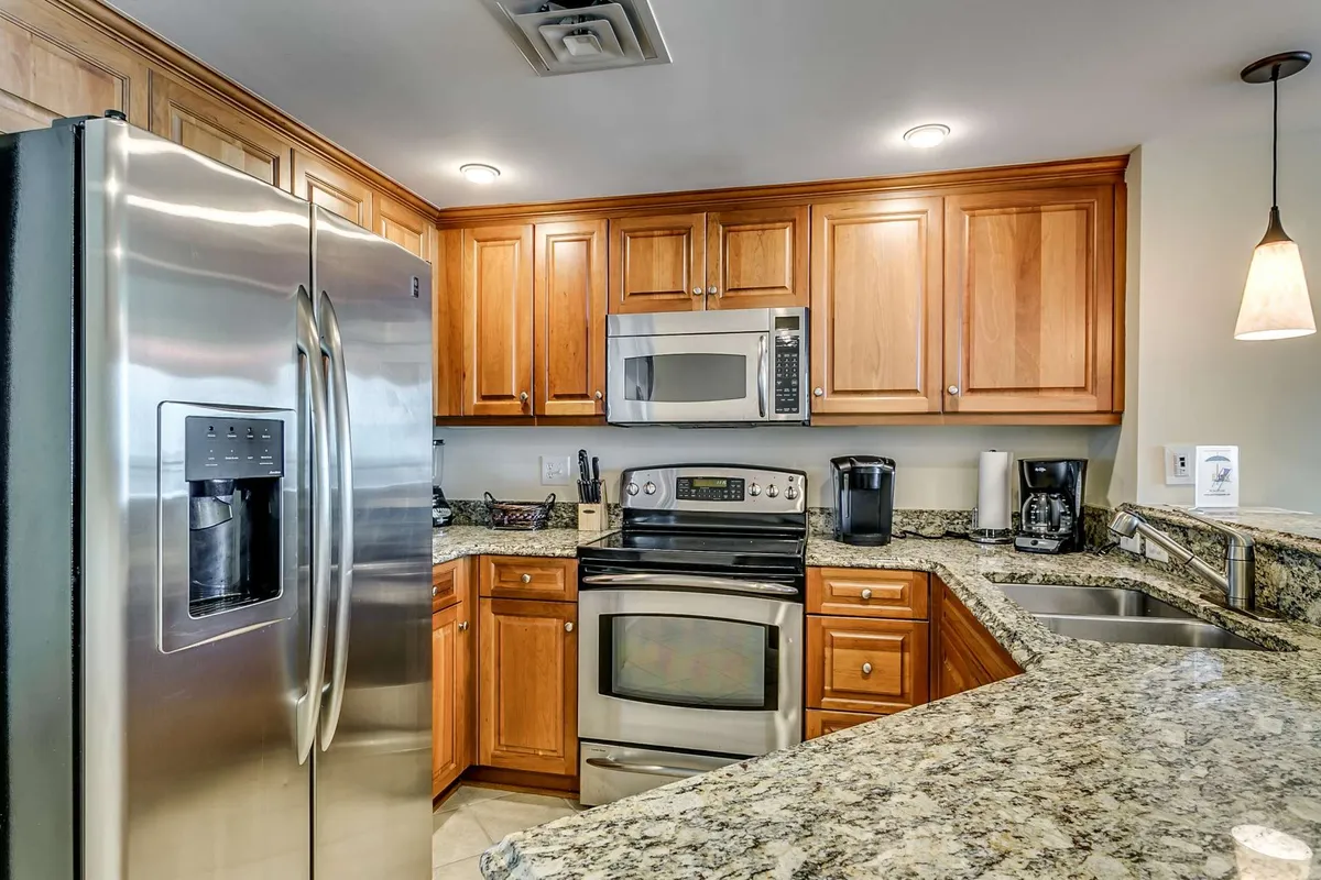 The image showcases a modern kitchen with stainless steel refrigerator, granite countertops, wooden cabinetry, and multiple kitchen appliances in view. The kitchen is spacious and well-lit, inviting for both cooking and entertaining.